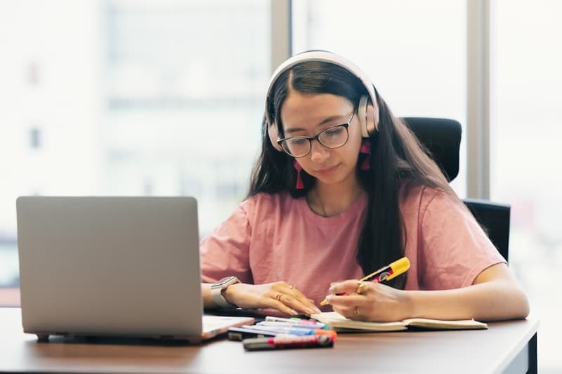 A student with headphones and glasses writing in her notebook with a laptop in front